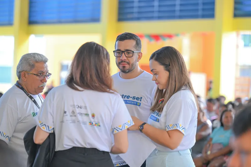 Integrantes das equipes, com camisetas do Registre-se! Brasil Parente, conversam durante os atendimentos; ao fundo, pessoas aguardam sentadas.